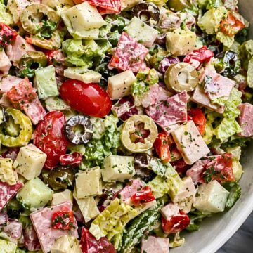 Antipasto grinder chopped salad with romaine lettuce, ham, salami, provolone, cherry tomatoes, cucumbers, artichoke hearts, roasted red peppers, banana peppers, black and green olives, and creamy dressing in a white bowl.