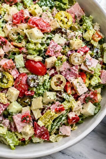 Antipasto grinder chopped salad with romaine lettuce, ham, salami, provolone, cherry tomatoes, cucumbers, artichoke hearts, roasted red peppers, banana peppers, black and green olives, and creamy dressing in a white bowl.