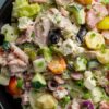 Close-up of antipasto grinder chopped salad with lettuce, deli meats, cucumbers, tomatoes, black olives, red onion, cheese, and creamy dressing in a black bowl.