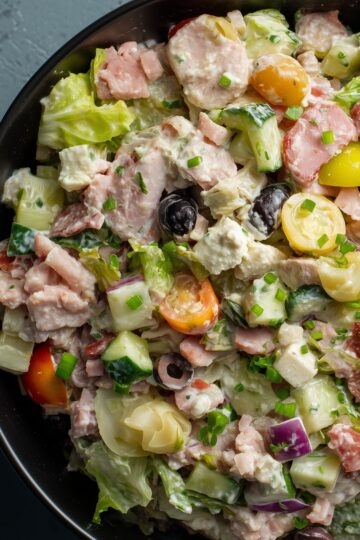 Close-up of antipasto grinder chopped salad with lettuce, deli meats, cucumbers, tomatoes, black olives, red onion, cheese, and creamy dressing in a black bowl.