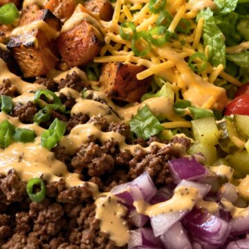 Cheeseburger sweet potato bowl with seasoned ground beef, roasted sweet potato cubes, romaine lettuce, shredded cheddar, pickles, red onion, green onions, tomatoes, and creamy burger sauce in a white bowl.