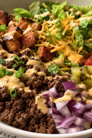 Cheeseburger sweet potato bowl with seasoned ground beef, roasted sweet potato cubes, romaine lettuce, shredded cheddar, pickles, red onion, green onions, tomatoes, and creamy burger sauce in a white bowl.
