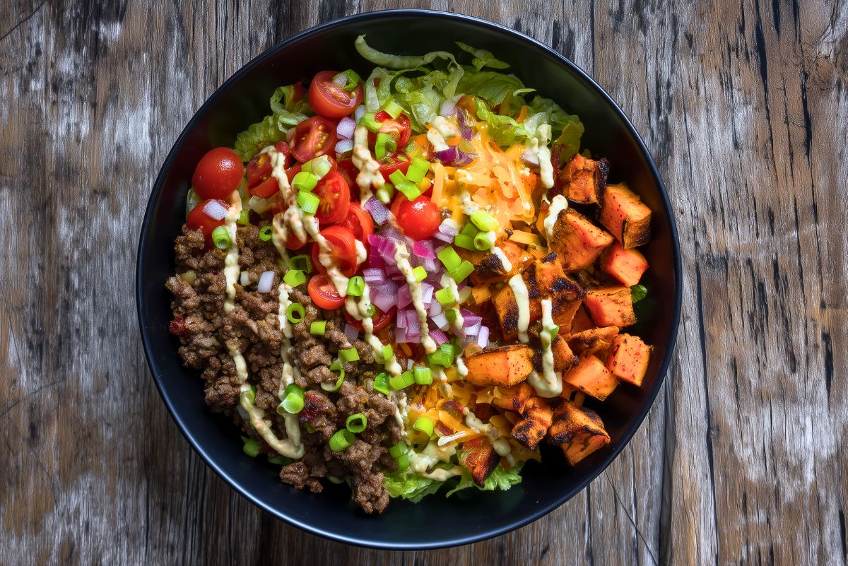 Cheeseburger sweet potato bowl with seasoned ground beef, roasted sweet potatoes, lettuce, cherry tomatoes, red onion, shredded cheddar, green onions, and creamy burger sauce.