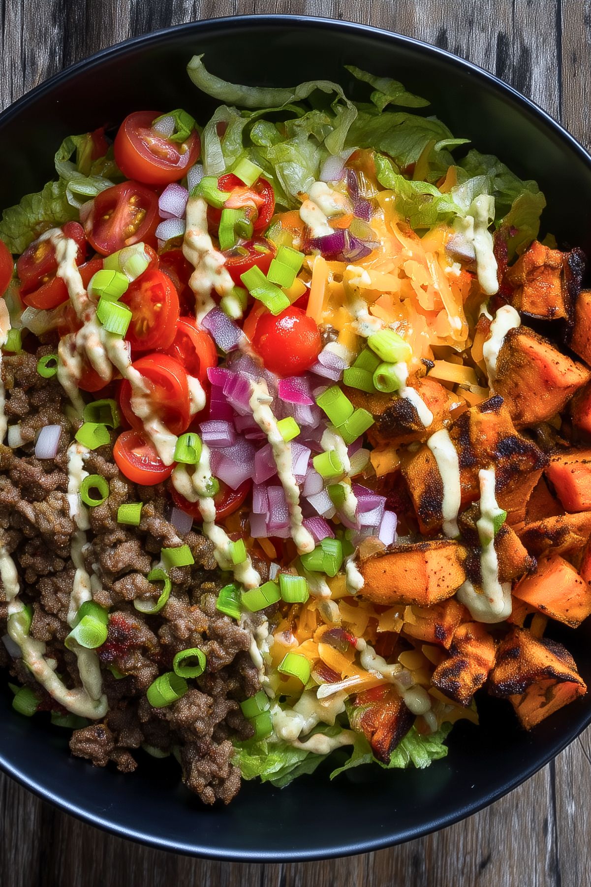 Cheeseburger sweet potato bowl with seasoned ground beef, roasted sweet potatoes, lettuce, cherry tomatoes, red onion, shredded cheddar, green onions, and creamy burger sauce.
