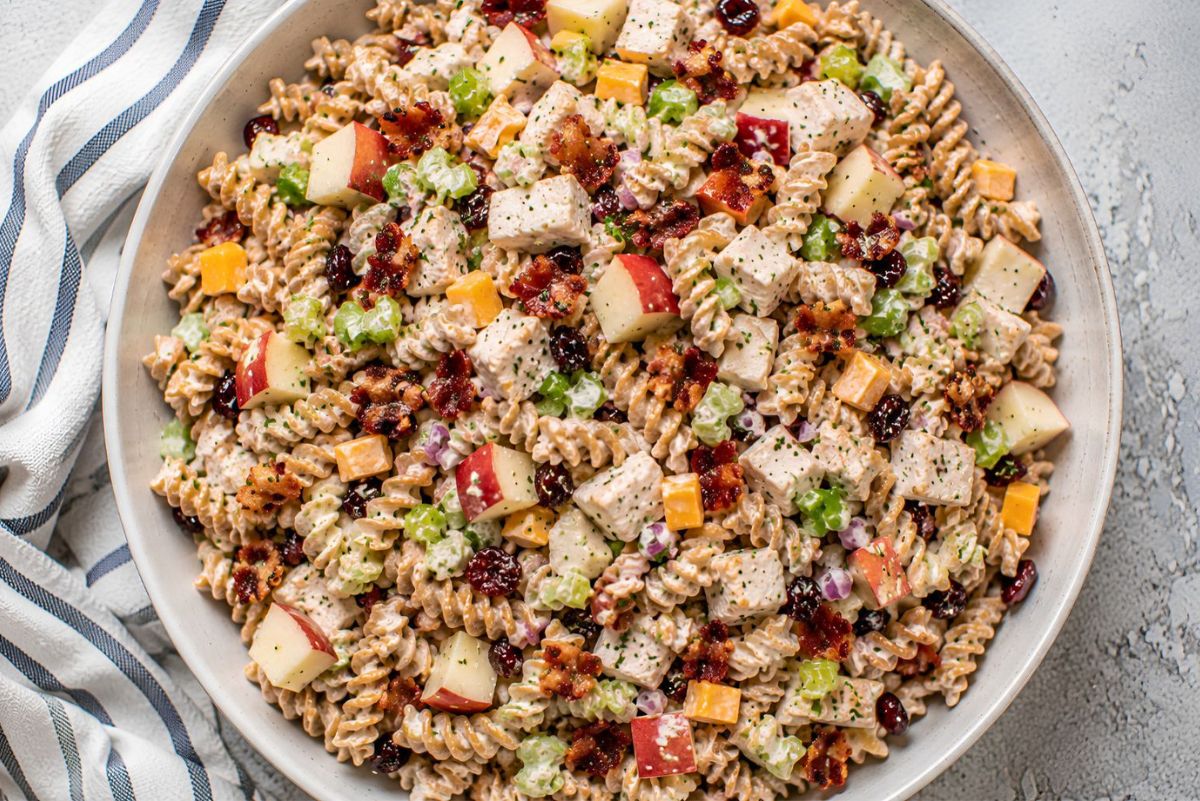 Chicken bacon apple pasta salad with whole wheat rotini, chopped chicken, crispy bacon, red apples, celery, cheddar cheese, dried cranberries, red onion, and creamy Dijon dressing in a large bowl.