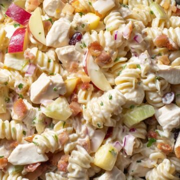 Close-up of creamy chicken bacon apple pasta salad with rotini, chicken, red apples, celery, red onion, cranberries, and crisp bacon in a black bowl.
