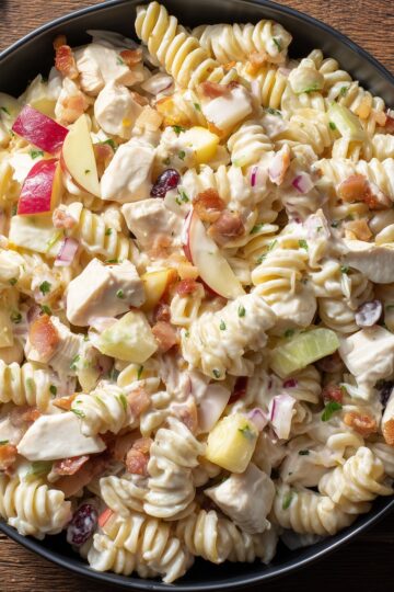Close-up of creamy chicken bacon apple pasta salad with rotini, chicken, red apples, celery, red onion, cranberries, and crisp bacon in a black bowl.