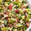 Close-up of club grinder chopped salad with romaine, turkey, ham, provolone, cherry tomatoes, cucumbers, black olives, banana peppers, bacon, herbs, and creamy dressing in a white bowl.