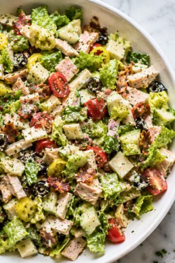 Close-up of club grinder chopped salad with romaine, turkey, ham, provolone, cherry tomatoes, cucumbers, black olives, banana peppers, bacon, herbs, and creamy dressing in a white bowl.