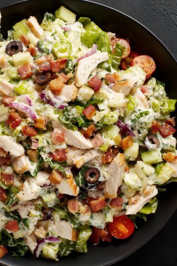 Close-up of club grinder chopped salad with turkey, bacon, lettuce, tomatoes, cucumbers, black olives, red onion, cheese, and creamy dressing in a black bowl.