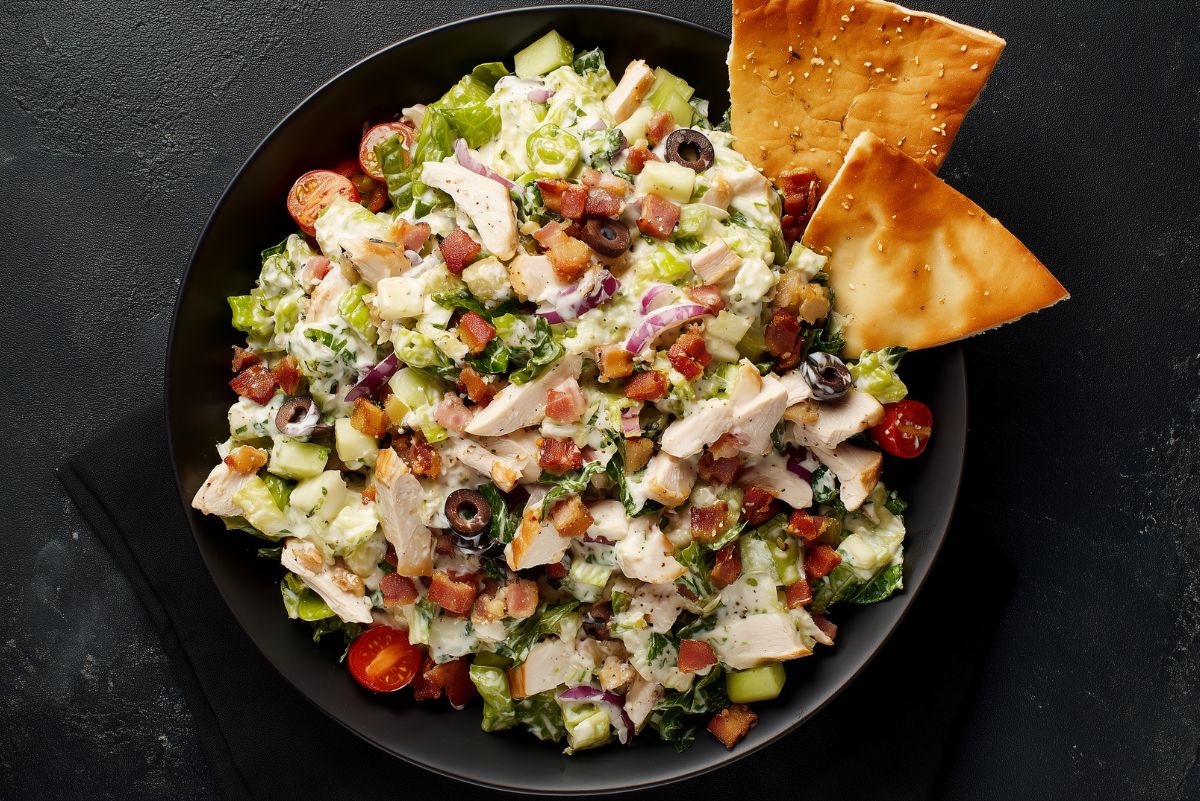 Overhead view of club grinder chopped salad with turkey, bacon, lettuce, tomatoes, cucumbers, black olives, red onion, cheese, and creamy dressing, served with pita chips.