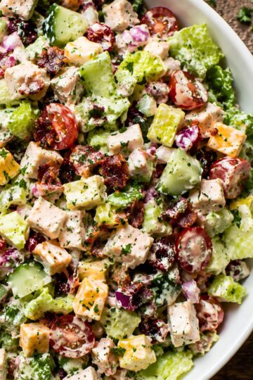 Close-up of cranberry turkey club chopped salad with chopped romaine, turkey breast, crispy bacon, cheddar cheese, cucumbers, cherry tomatoes, red onion, dried cranberries, and creamy dressing.