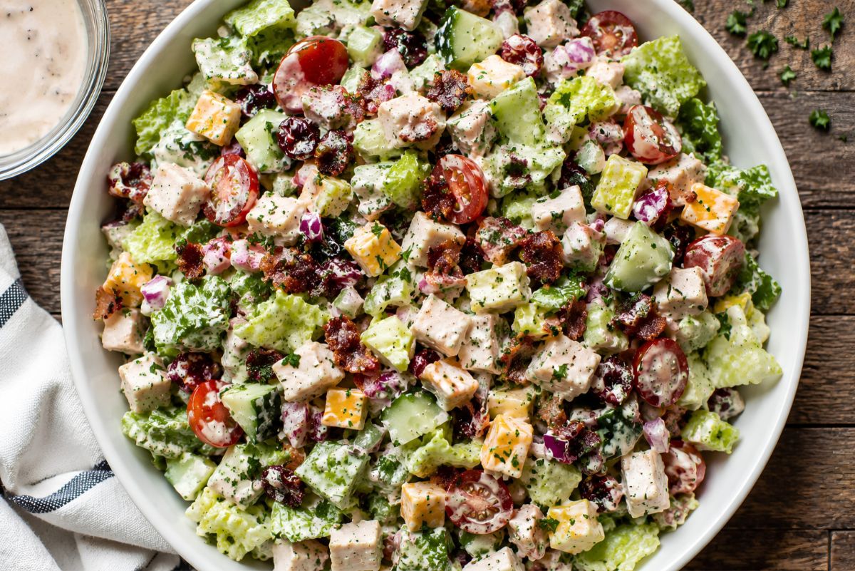 Overhead view of cranberry turkey club chopped salad with romaine, turkey breast, bacon, cheddar, cucumbers, cherry tomatoes, red onion, dried cranberries, and creamy dressing, served with extra dressing on the side.