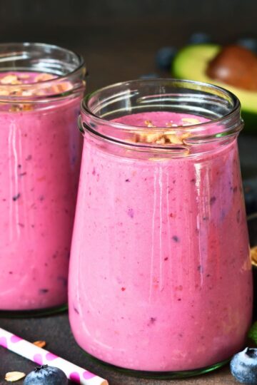 Creamy berry avocado smoothie in glass jars with a pink fruit blend, topped with oats or granola, with blueberries, a striped straw, and sliced avocado in the background.