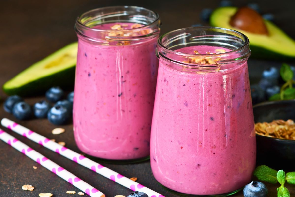 Two creamy berry avocado smoothies in glass jars with bright pink berry color, topped with oats or granola, with blueberries, avocado halves, mint, granola, and pink striped straws on a dark tabletop.