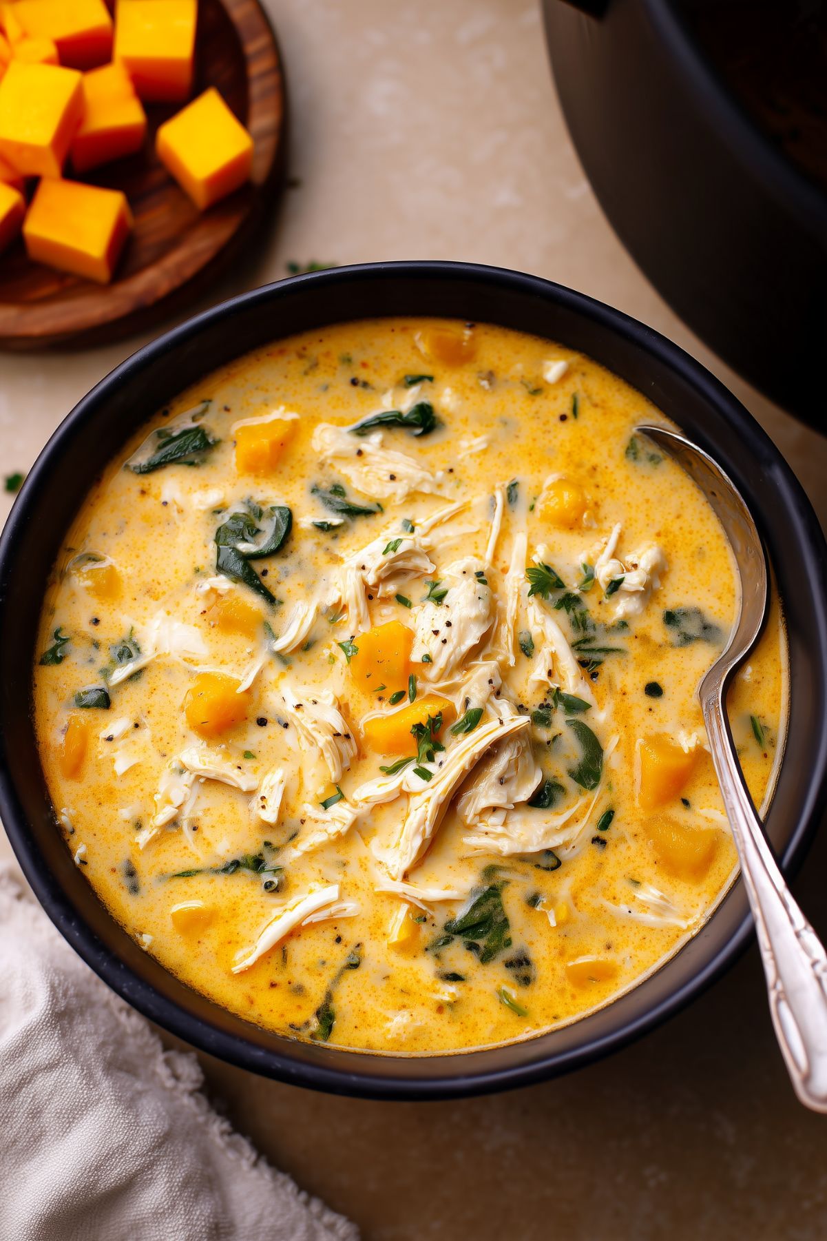 Creamy chicken and butternut squash soup in a black bowl with shredded chicken, tender squash cubes, leafy greens, herbs, and a rich golden broth. A spoon rests in the bowl, with cubed butternut squash in the background.