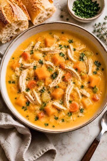 Creamy chicken and butternut squash soup with shredded chicken, squash cubes, carrots, celery, parsley, and black pepper in a rustic bowl, served with crusty bread.