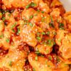 Crockpot honey orange chicken coated in a glossy orange sauce and topped with sesame seeds and chopped chives in a white serving bowl.
