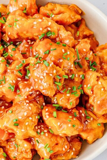 Crockpot honey orange chicken coated in a glossy orange sauce and topped with sesame seeds and chopped chives in a white serving bowl.
