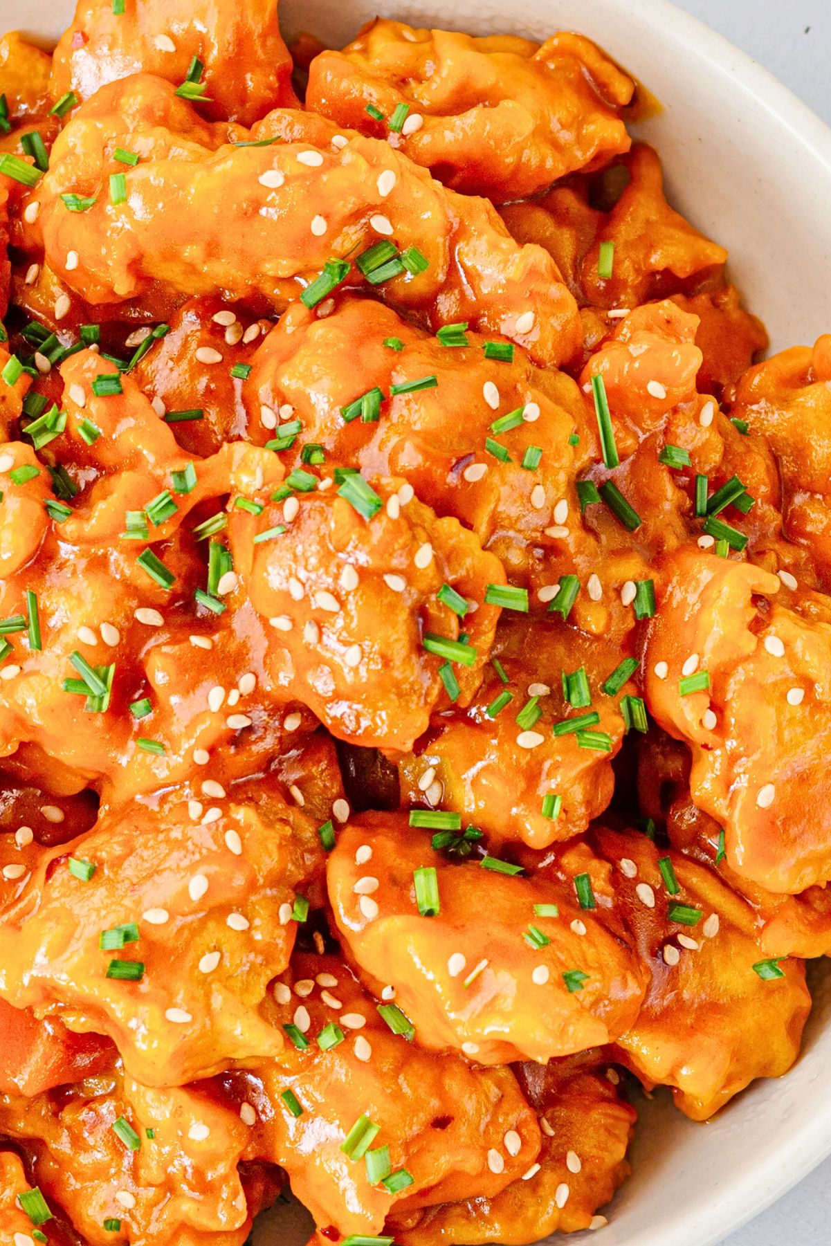 Crockpot honey orange chicken coated in a glossy orange sauce and topped with sesame seeds and chopped chives in a white serving bowl.