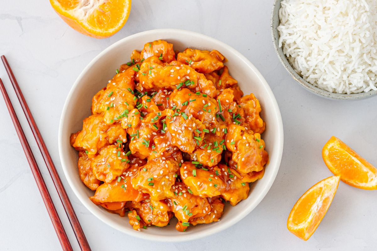 Crockpot honey orange chicken topped with sesame seeds and chopped chives, served beside white rice, orange wedges, and red chopsticks.