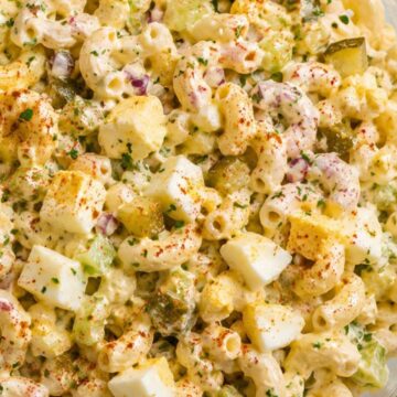 Close-up of creamy deviled egg pasta salad with elbow macaroni, chopped hard-boiled eggs, celery, dill pickles, red onion, fresh herbs, and paprika in a glass bowl.