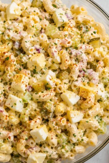 Close-up of creamy deviled egg pasta salad with elbow macaroni, chopped hard-boiled eggs, celery, dill pickles, red onion, fresh herbs, and paprika in a glass bowl.