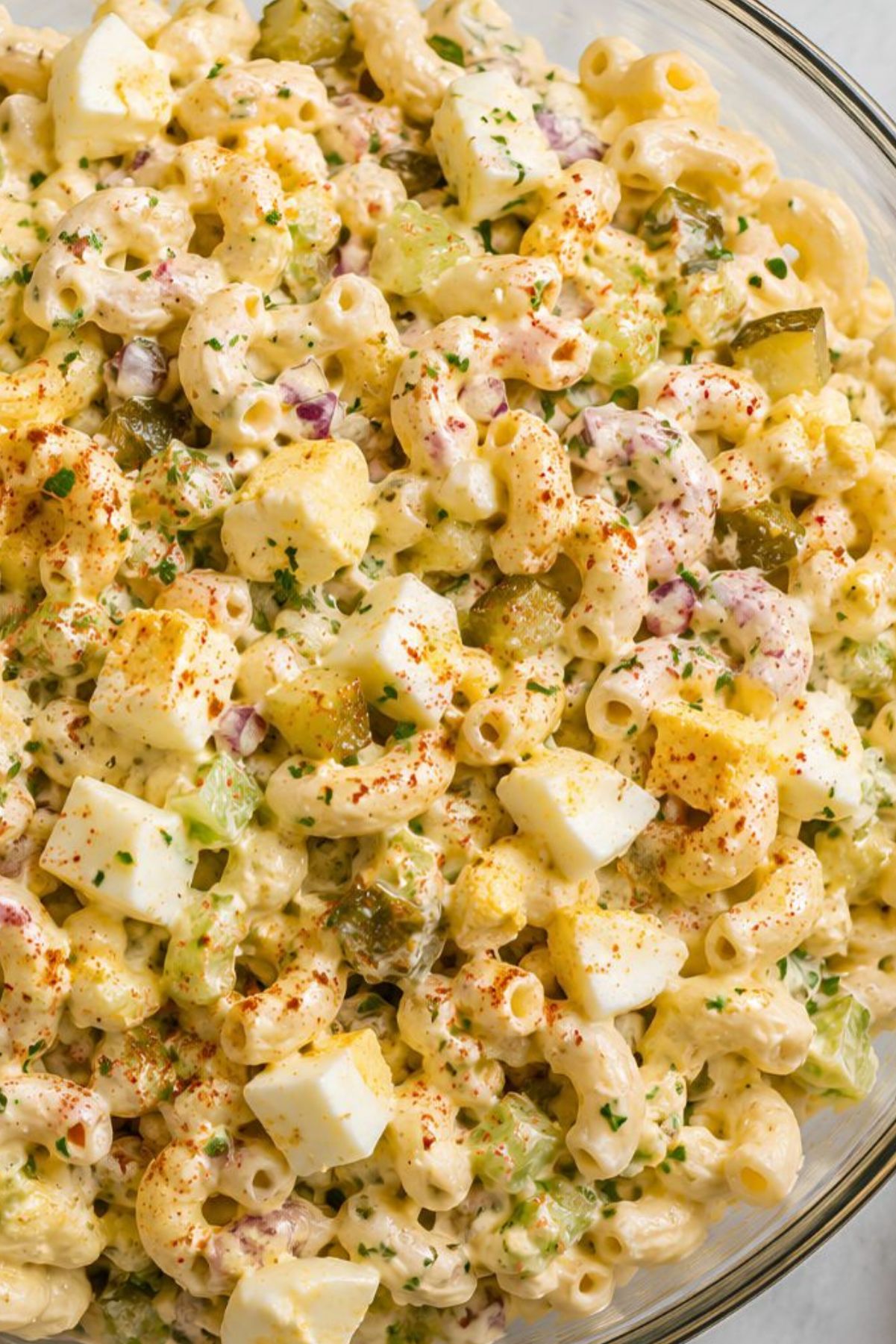 Close-up of creamy deviled egg pasta salad with elbow macaroni, chopped hard-boiled eggs, celery, dill pickles, red onion, fresh herbs, and paprika in a glass bowl.