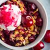Microwave cherry crisp in a small bowl with glossy cherries, golden oat crumble, and a scoop of whipped cream swirled with cherry sauce. Fresh cherries sit on the plate beside the warm dessert.