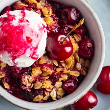 Microwave cherry crisp in a small bowl with glossy cherries, golden oat crumble, and a scoop of whipped cream swirled with cherry sauce. Fresh cherries sit on the plate beside the warm dessert.