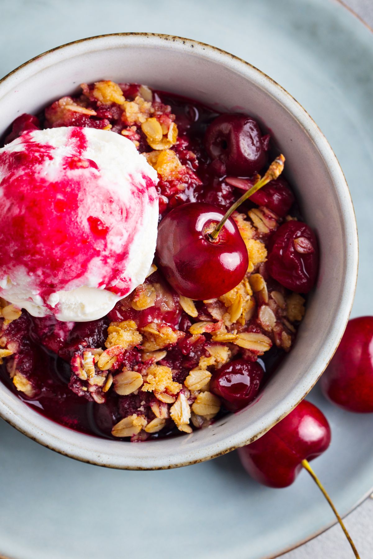 Microwave cherry crisp in a small bowl with glossy cherries, golden oat crumble, and a scoop of whipped cream swirled with cherry sauce. Fresh cherries sit on the plate beside the warm dessert.
