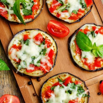 Eggplant Margherita pizza bites made with roasted eggplant rounds topped with melted mozzarella, chopped tomatoes, and fresh basil, arranged on a wooden cutting board with tomato wedges and basil leaves.