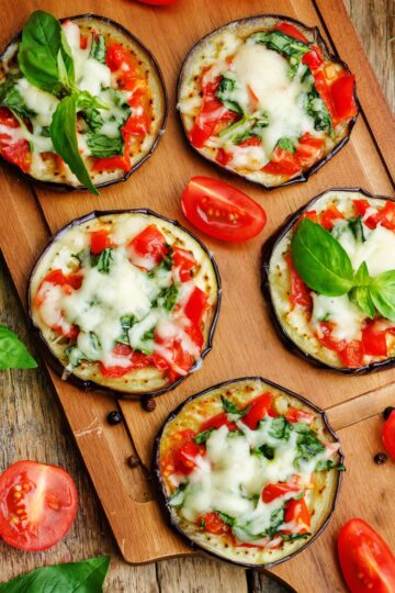 Eggplant Margherita pizza bites made with roasted eggplant rounds topped with melted mozzarella, chopped tomatoes, and fresh basil, arranged on a wooden cutting board with tomato wedges and basil leaves.