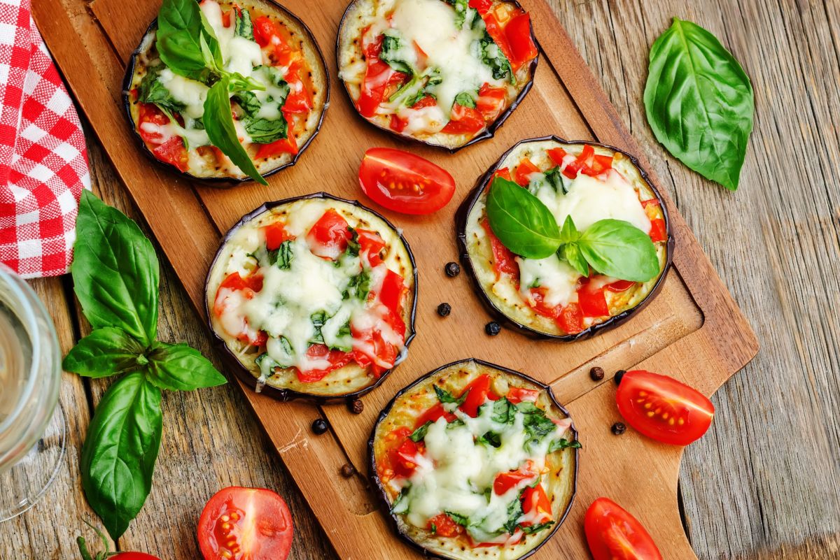 Eggplant Margherita pizza bites topped with melted mozzarella, diced tomatoes, and fresh basil on a wooden cutting board, surrounded by tomato wedges, basil leaves, peppercorns, and a red-and-white checkered napkin.