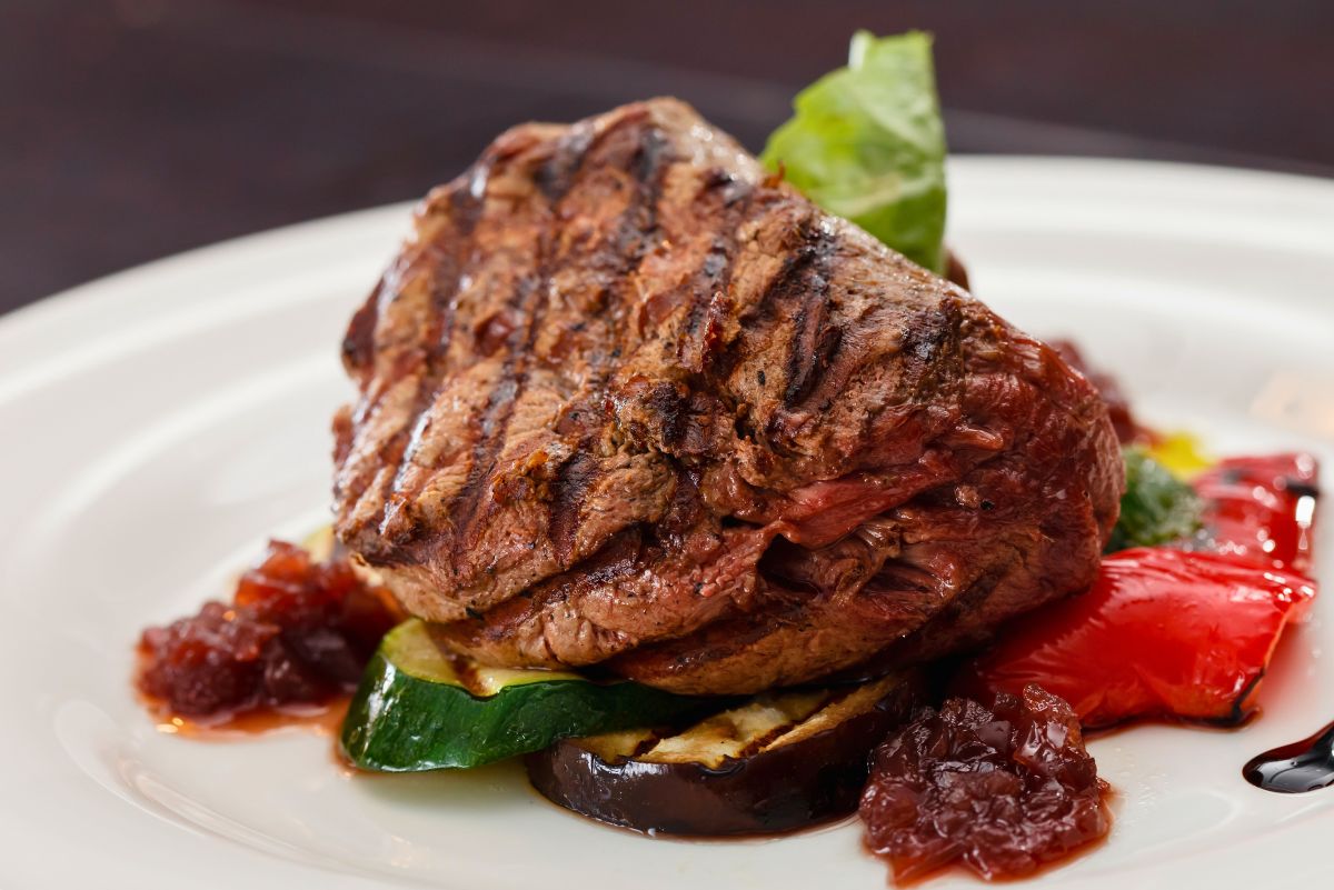 Grilled sirloin steak with charred grill marks served over zucchini and eggplant, with roasted red pepper, balsamic glaze, and chunky tomato-onion jam on a white plate.