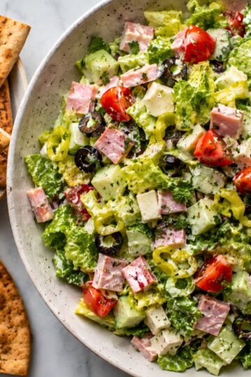 Creamy ham and provolone grinder chopped salad with romaine lettuce, cucumber, cherry tomatoes, banana peppers, black olives, herbs, and garlic pita chips served on the side.