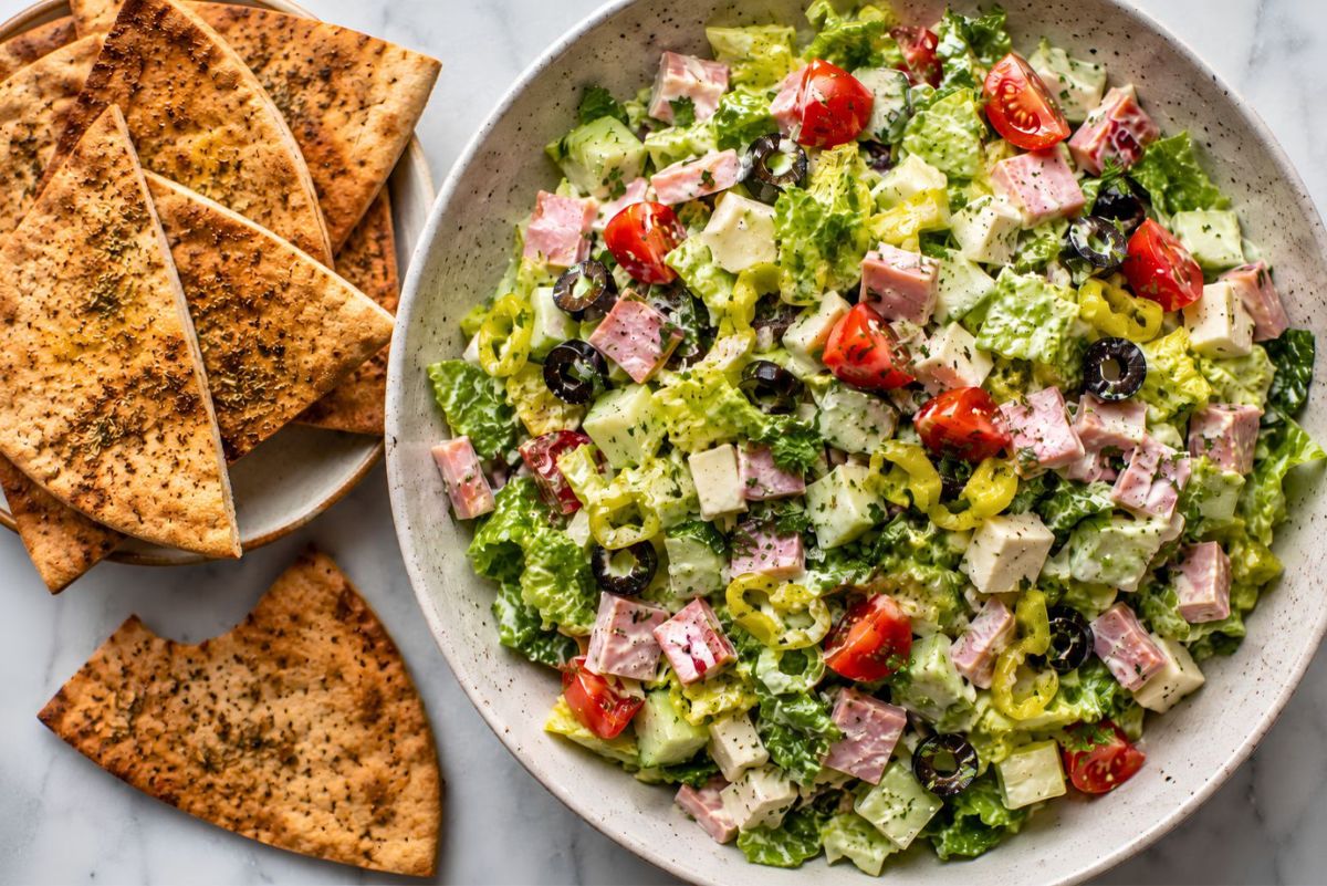 Ham and provolone grinder chopped salad with romaine, cucumber, cherry tomatoes, banana peppers, black olives, creamy dressing, and seasoned garlic pita chips on the side.