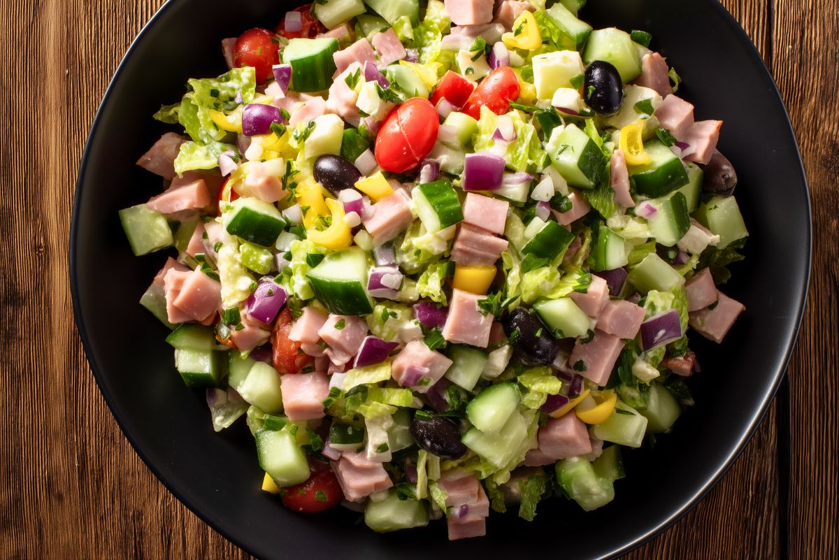 Ham and provolone grinder chopped salad with diced ham, provolone, lettuce, cucumbers, tomatoes, red onion, banana peppers, black olives, and creamy dressing in a black bowl.