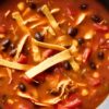 Hearty chicken tortilla soup with shredded chicken, black beans, corn, tomatoes, and crispy tortilla strips in a black bowl.