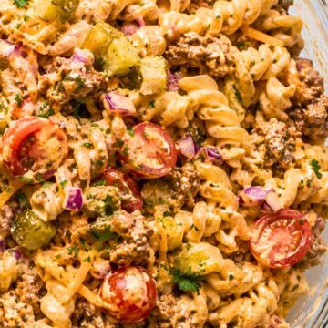 High protein cheeseburger pasta salad with rotini, seasoned ground beef, cheddar cheese, cherry tomatoes, dill pickles, red onion, parsley, and creamy cheeseburger dressing in a glass serving bowl.