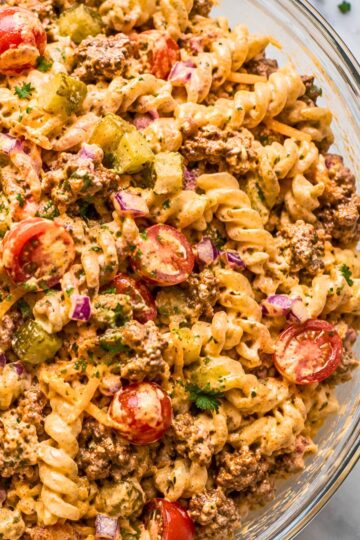 High protein cheeseburger pasta salad with rotini, seasoned ground beef, cheddar cheese, cherry tomatoes, dill pickles, red onion, parsley, and creamy cheeseburger dressing in a glass serving bowl.