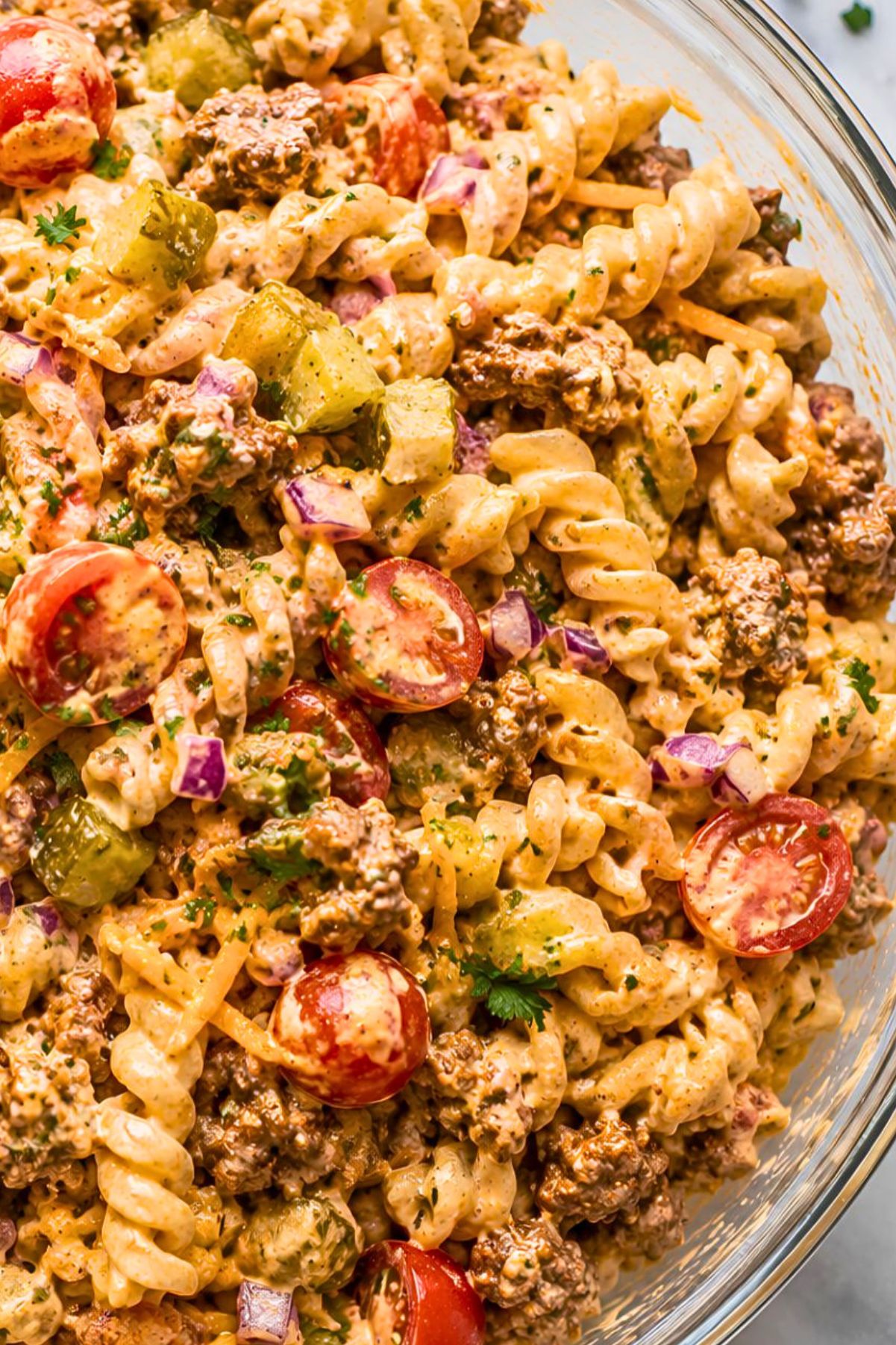 High protein cheeseburger pasta salad with rotini, seasoned ground beef, cheddar cheese, cherry tomatoes, dill pickles, red onion, parsley, and creamy cheeseburger dressing in a glass serving bowl.