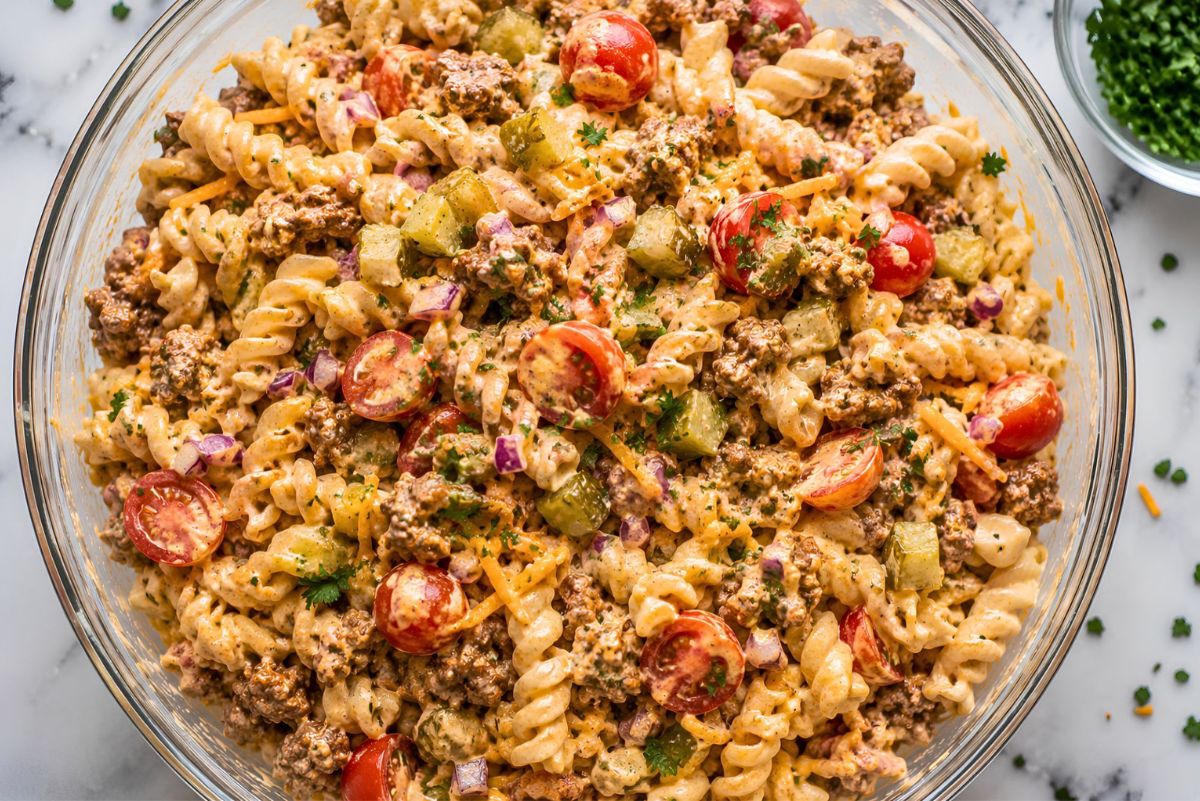High protein cheeseburger pasta salad with rotini, seasoned ground beef, shredded cheddar, cherry tomatoes, dill pickles, red onion, parsley, and creamy dressing in a large glass bowl.