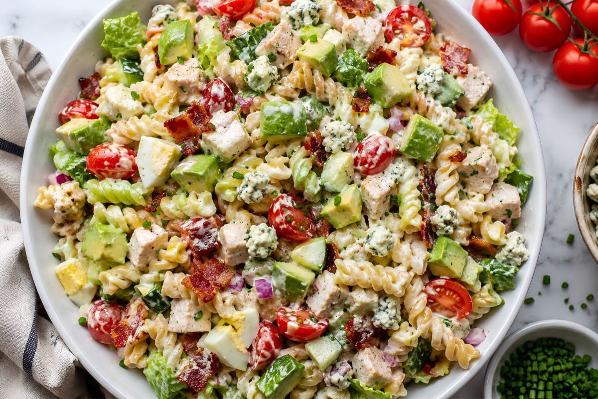 High protein Chicken Cobb pasta salad with rotini, chicken breast, turkey bacon, avocado, cherry tomatoes, cucumber, romaine, hard-boiled eggs, red onion, blue cheese crumbles, and creamy Cobb dressing.