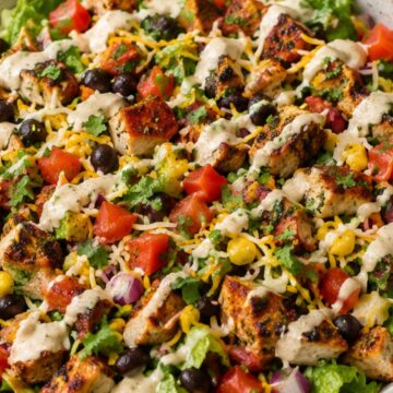High protein chicken taco salad bowl with grilled chicken, romaine lettuce, black beans, corn, tomatoes, red onion, shredded cheese, cilantro, and creamy taco dressing in a white bowl.