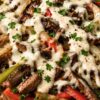 High protein Philly cheesesteak bowl with sliced steak, red and green bell peppers, onions, mushrooms, brown rice, melted provolone cheese, creamy sauce, black pepper, and fresh parsley in a white bowl.