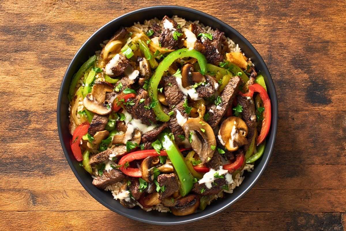 High protein Philly cheesesteak bowl with sliced steak, rice, sautéed mushrooms, green and red bell peppers, onions, creamy cheese sauce, and fresh parsley.