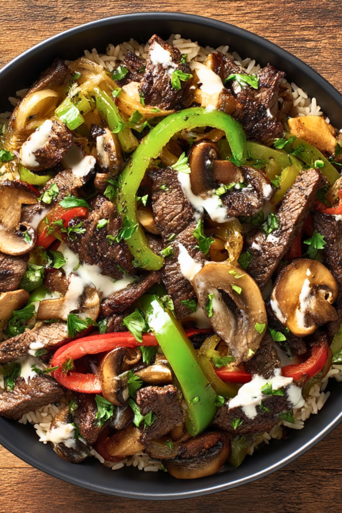 High protein Philly cheesesteak bowl with sliced steak, rice, sautéed mushrooms, bell peppers, onions, creamy cheese sauce, and fresh parsley in a black bowl.