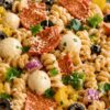 High protein pizza pasta salad with creamy rotini, quartered turkey pepperoni, mozzarella pearls, black olives, green bell pepper, banana peppers, red onion, Parmesan, and fresh parsley in a clear glass bowl.