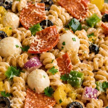 High protein pizza pasta salad with creamy rotini, quartered turkey pepperoni, mozzarella pearls, black olives, green bell pepper, banana peppers, red onion, Parmesan, and fresh parsley in a clear glass bowl.